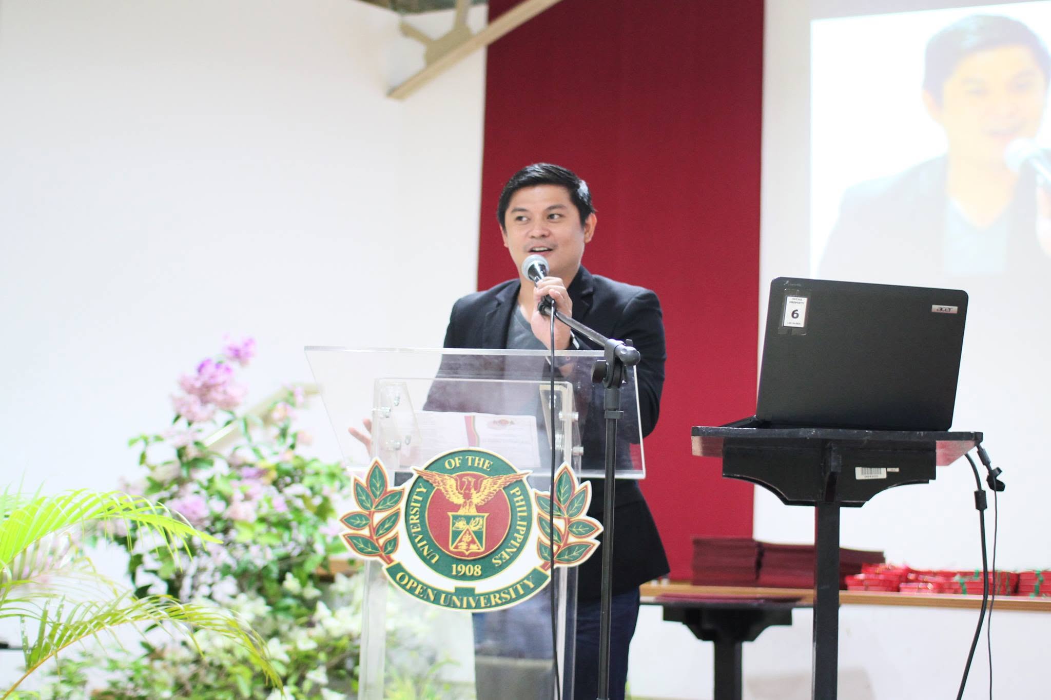 About 3 RM Nisperos speaking at a lectern bearing the University of the Philippines Open University seal during a lecture
